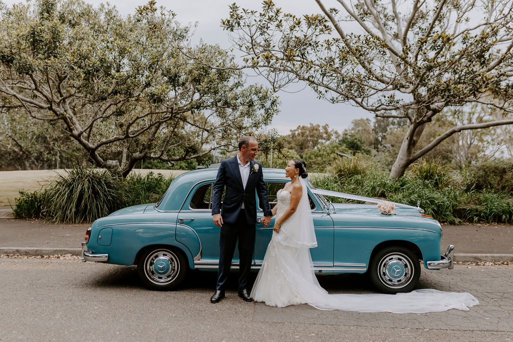 Bride and groom holding hands next to a classic blue Mercedes-Benz Ponton wedding car in a lush garden setting.