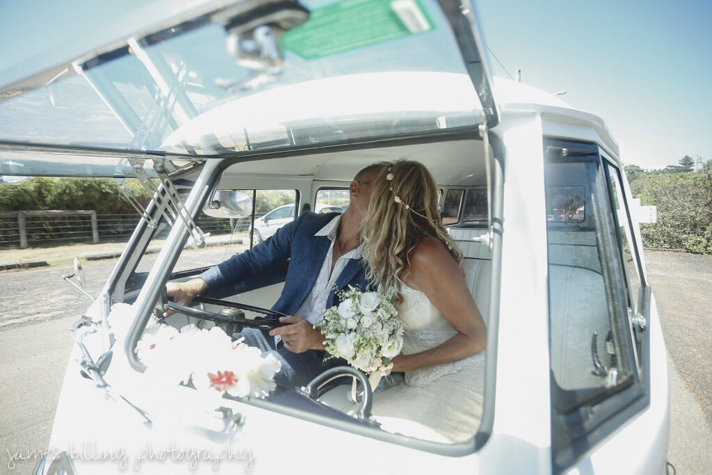 Bride kissing groom staged in the driver's seat through the open front safari window of a vintage white Kombi van.