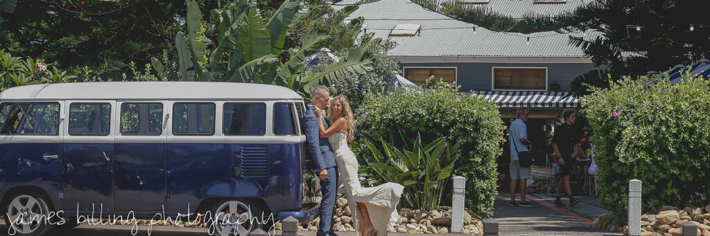 Bride and groom posing next to a vintage VW Kombi wedding car at a beachside venue.